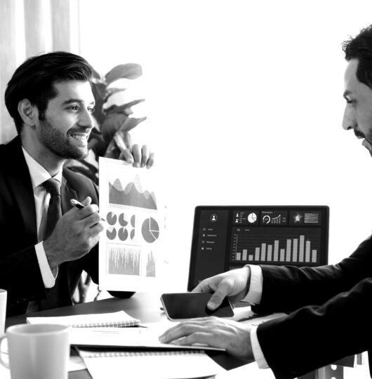 two-men-suits-sit-desk-one-them-is-holding-graph-other-is-showing-graph-scaled-blackwhite